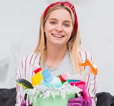 A smiling person wearing gloves holds a variety of cleaning products.