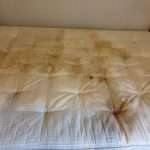 A close-up photo of a heavily soiled and stained mattress, left behind by tenants who have moved out. The mattress appears to be old and worn, with visible dirt, dust, and stains covering its surface. The bedding is in disarray, and there are visible tears and holes in the fabric