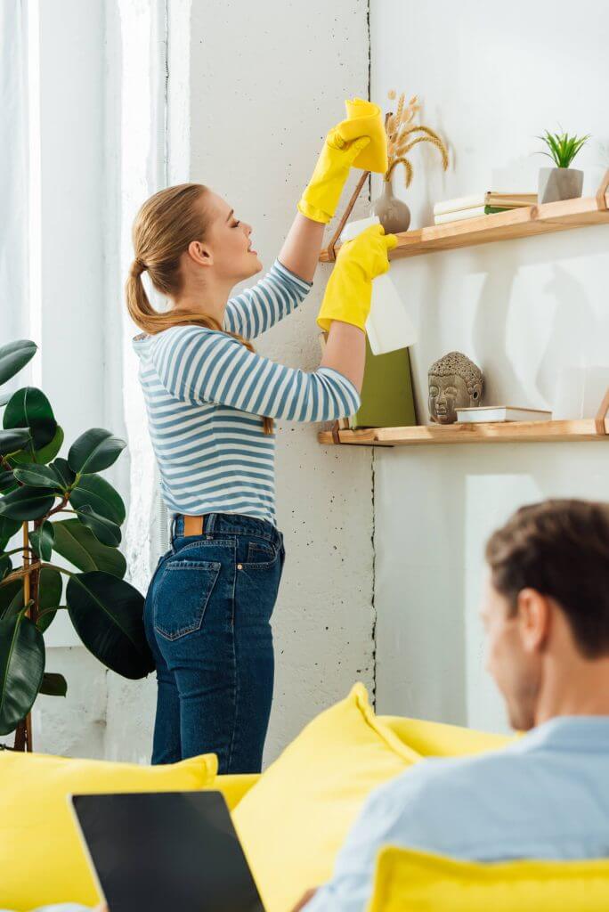 Woman in uniform cleaning kitchen countertop with spray and cloth.